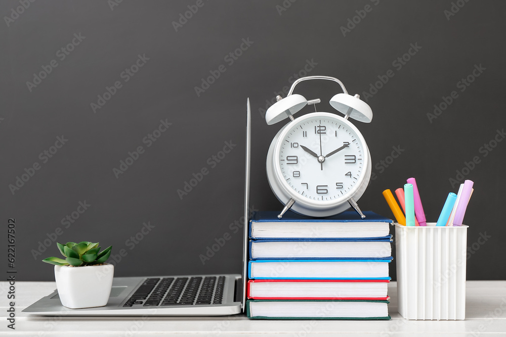 Different stationery with alarm clock and laptop on white table against black chalkboard
