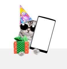 Poster - Cute kitten wearing sunglasses and party cap holds gift box and shows big smartphone with blank screen above empty white banner. isolated on white background