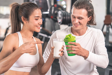 Healthy lifestyle and healthy food concept. Asian woman wearing sports bra holding glass of milk compliments Caucasian male trainer's whey protein glass. Both finish exercising drinking healthy drink 