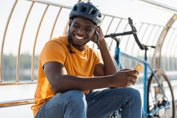 Authentic portrait of smiling African American man wearing protective helmet holding mobile phone, communication online sitting near modern bicycle on the street. Hobby, positive lifestyle concept