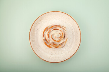 Cinnamon roll or cinnabon in paper box on the plate on green background, top view. Homemade sweet traditional dessert buns with white cream sauce