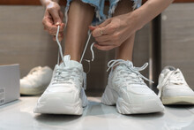 Close-up Of Women's Legs Choosing Sports Shoes And Trying On Different White Sneakers At The Mall. Women's Hands Close-up Tie Shoelaces When Trying On Sports Shoes.