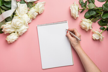 Female hands writing in notebook and bouquet white roses on pink background. Top view. Copy space.