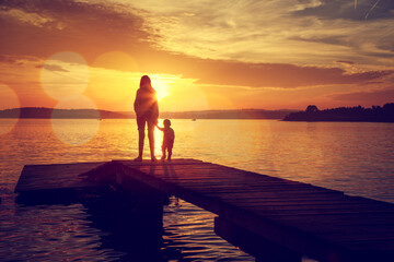 Poster - Silhouettes of Young Mother and Her Little Baby Standing on the Pier and Watching Sunset by the Lake. Happy Family Lifestyle. Motherhood and Childhood. Mom and Son. Toned Photo with Bokeh. Copy Space.