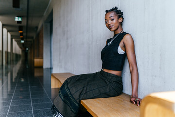 Portriat of african millennial woman posing indoor sitting on bench over gray wall background. Female student sitting in hallway interior at campus while resting after long hard lessons.