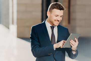 Succesful male entrepreneur in formal suit, holds modern tablet, checks documentation online, reads necessary information for making business report, uses free wifi connection. People and technology