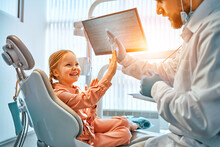 A Little Girl Is Sitting In A Dentist's Chair, Giving A High Five To The Doctor And Laughing. Dental Care, Trust And Patient Care. Children's Dentistry.Sunlight.