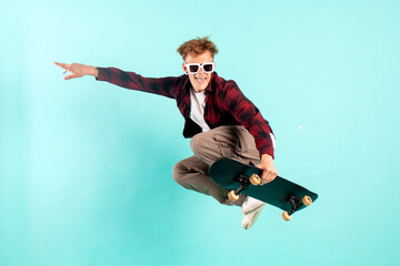 Wall Mural - young crazy guy rides skateboard and jumps on blue isolated background, hipster in sunglasses flies