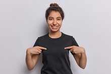 Horizontal Shot Of Pretty Iranian Girl Points Fingers At Blank Copy Space Of T Shirt Smiles Pleasantly Suggests To Place Your Logo Or Design Here Isolated Over White Background. Clothing Template