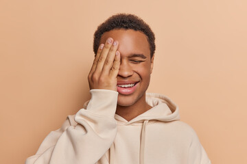 Wall Mural - Portrait of attractive man has dark skin hides half of face with palm while smilies expresses friendliness. Guy shows only positive side of his temper dressed in casual hoodie isolated over brown wall