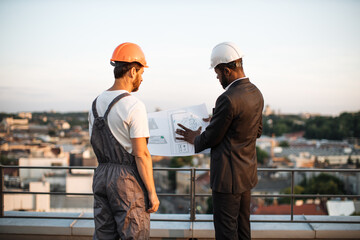 Rare view of caucasian bearded workman presenting design blueprints to african american urban planner on fresh air. Male developers in hardhats discussing location of balconies on apartment building.