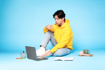 Wall Mural - Thoughtful korean teenager guy using laptop sitting on blue background