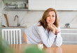 Woman in unbuttoned white shirt is standing in kitchen and leaning elbows on dining table. Blurred kitchen utensil dishes in background. Sensual professional shot