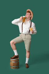 Young man in traditional German clothes with beer and pretzel on green background