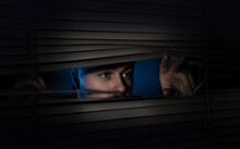 Worried Man Looking Through Window Blinds Into Darkness. Paranoia Concept