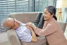 Asian Senior Man Lying Down And Suffering From Chest Pain Or Heart Attack From Working Accident On Sofa In Living Room. Elderly Woman Caregiver Consoling And Help While Hugging Support And First Aid