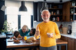 Happy positive senior man looking at camera and smiling showing thumb up as sign of approval satisfied with breakfast cereals, standing in front of members of family, senior woman and two kids.
