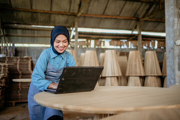 Wall Mural - female Muslim entrepreneur wearing apron using laptop computer on wooden table in wood craft shop