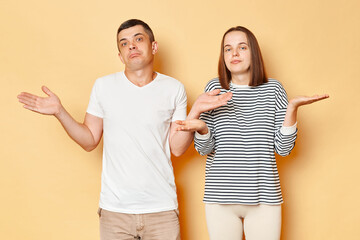 Wall Mural - Uncertain puzzled young couple wife and husband wearing casual attires standing isolated over beige background shrugging shoulders don't know what to say.