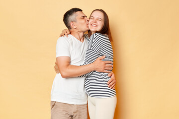 Wall Mural - Loving young couple wife and husband wearing casual attires standing isolated over beige background hugging each other and man kissing his girlfiend with happy face.