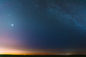 Wall Mural - Night Starry Sky With Glowing Stars Above Countryside Field Landscape In Early Spring. Bright Glow Of Planet Venus In Sky Among The Stars. Sky In Warm Lights Of Evening Sunset Dawn Or Morning Sunrise.