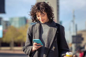 Wall Mural - Young woman in coat using mobile phone while walking outdoors in city