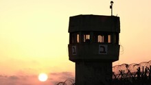 A Silhouette Of A Prison Watchtower At Sunset. The Sun Ball Is Beautifully Set At The Bottom Of The Frame, While Some Barbed Wire Is Apparent As Well.
