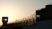 A Silhouette Of A Prison Watchtower At Sunset. The Barbed Wire Is Apparent At The Bottom Of The Frame And The Sun Ball Itself Is Visible Directly Through The Tower Windows.