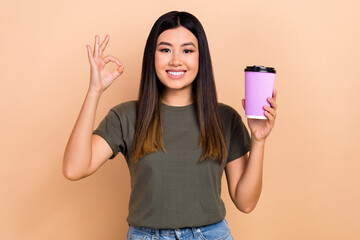 Wall Mural - Photo of cute cheerful thai girl toothy smile hand hold coffee cup demonstrate okey symbol isolated on beige color background