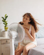 © MyJuly - Caucasian woman cools down at home with help of an air conditioner fan, sits on bed in front of an electric ventilator during intense heat of house, heat stroke, malaise from temperature in summer
