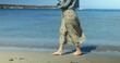 Stylish student girl strolling on wet beach sand along coastline on sunny day. Anonymous woman in long skirt and denim jacket walks with bare feet on shore in water.