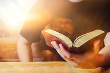 Close Up Of A Man Hand Holding The Holy Bible On Wooden Table Against The Sunlight With Bokeh, Christian Bible Study Or Devotional Concept Copy Space For Your Text