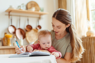 Young beautiful mother holding little cute baby working together from home, writing in notebook