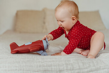 Wall Mural - Portrait of happy positive baby sitting on bed in living room at home, playing with toy airplane