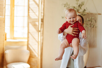 Wall Mural - Beautiful mother holding and playing with little cute baby in living room at home, copy space