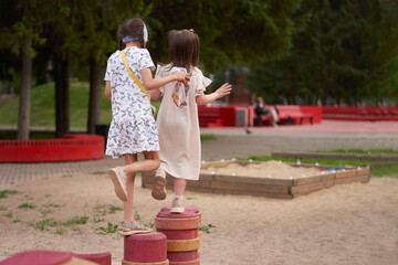 The girls are playing on a street playground. They keep balance standing on one leg on vertical poles. In the background is the entourage of the neighborhood.