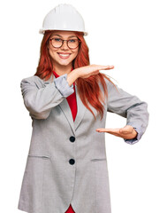 Poster - Young redhead woman wearing architect hardhat gesturing with hands showing big and large size sign, measure symbol. smiling looking at the camera. measuring concept.