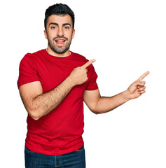 Wall Mural - Hispanic man with beard wearing casual red t shirt smiling and looking at the camera pointing with two hands and fingers to the side.