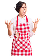 Poster - Beautiful young woman with short hair wearing professional cook apron crazy and mad shouting and yelling with aggressive expression and arms raised. frustration concept.