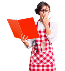 Canvas Print - Beautiful young woman with short hair wearing professional baker apron reading cooking recipe book covering mouth with hand, shocked and afraid for mistake. surprised expression