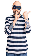 Wall Mural - Young handsome man wearing burglar mask asking to be quiet with finger on lips pointing with hand to the side. silence and secret concept.