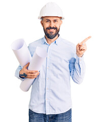 Canvas Print - Young handsome man wearing hardhat holding paper blueprints smiling happy pointing with hand and finger to the side