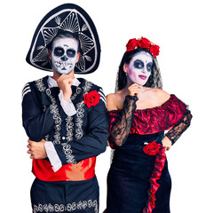 Poster - Young couple wearing mexican day of the dead costume over background looking confident at the camera smiling with crossed arms and hand raised on chin. thinking positive.