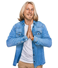 Poster - Caucasian man with blond long hair wearing casual denim jacket praying with hands together asking for forgiveness smiling confident.