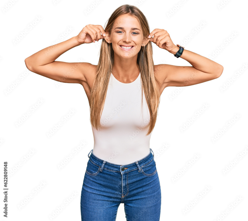 Young blonde woman wearing casual style with sleeveless shirt smiling pulling ears with fingers, funny gesture. audition problem