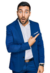 Poster - Young hispanic man wearing business jacket surprised pointing with finger to the side, open mouth amazed expression.