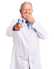 Canvas Print - Senior handsome grey-haired man wearing doctor coat and stethoscope laughing at you, pointing finger to the camera with hand over mouth, shame expression