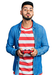 Wall Mural - Young hispanic man with beard playing video game holding controller looking at the camera blowing a kiss being lovely and sexy. love expression.