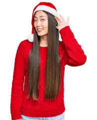 Poster - Young chinese woman wearing christmas hat smiling with hand over ear listening an hearing to rumor or gossip. deafness concept.