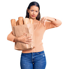 Sticker - Young beautiful latin girl holding paper bag with bread with angry face, negative sign showing dislike with thumbs down, rejection concept